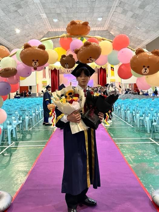 Shaun in a graduation gown and cap, holding two bouquets